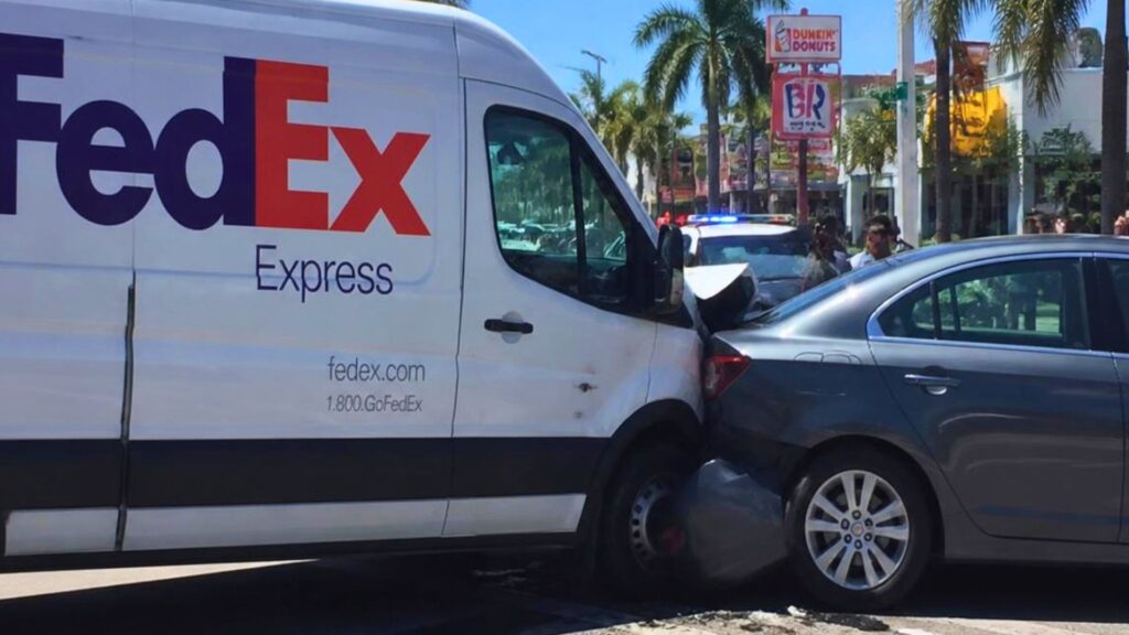 Dramatic side angle view of a FedEx Express delivery van crashing into a sedan with glass and debris flying along a palm tree lined Florida roadway.