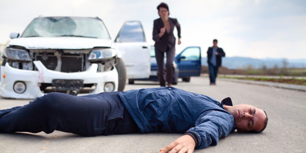 Injured pedestrian lying on the road in front of a damaged car while a witness rushes to help after a vehicle collision.